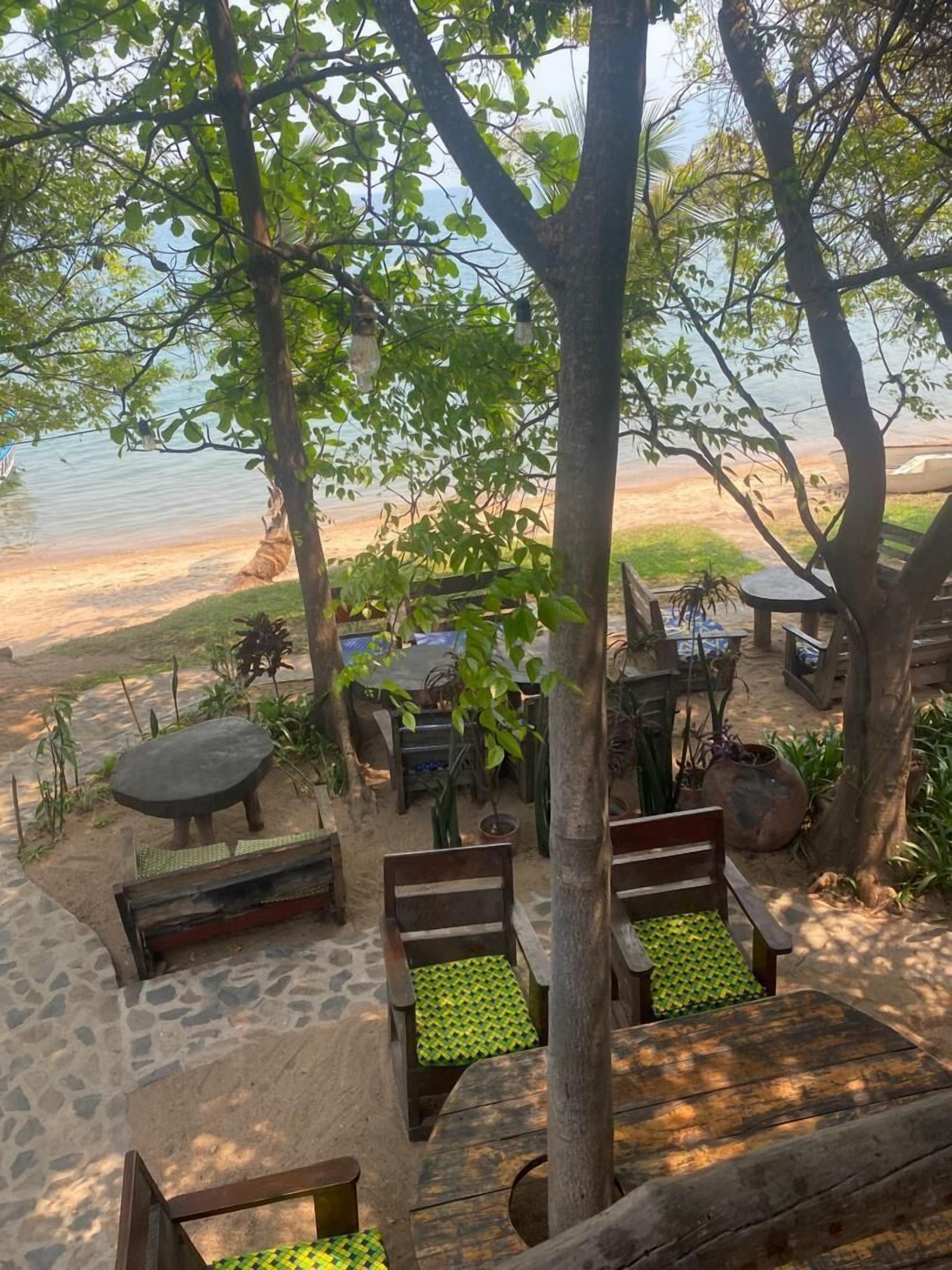 A beachside seating area with wooden benches and tables, surrounded by trees and plants.