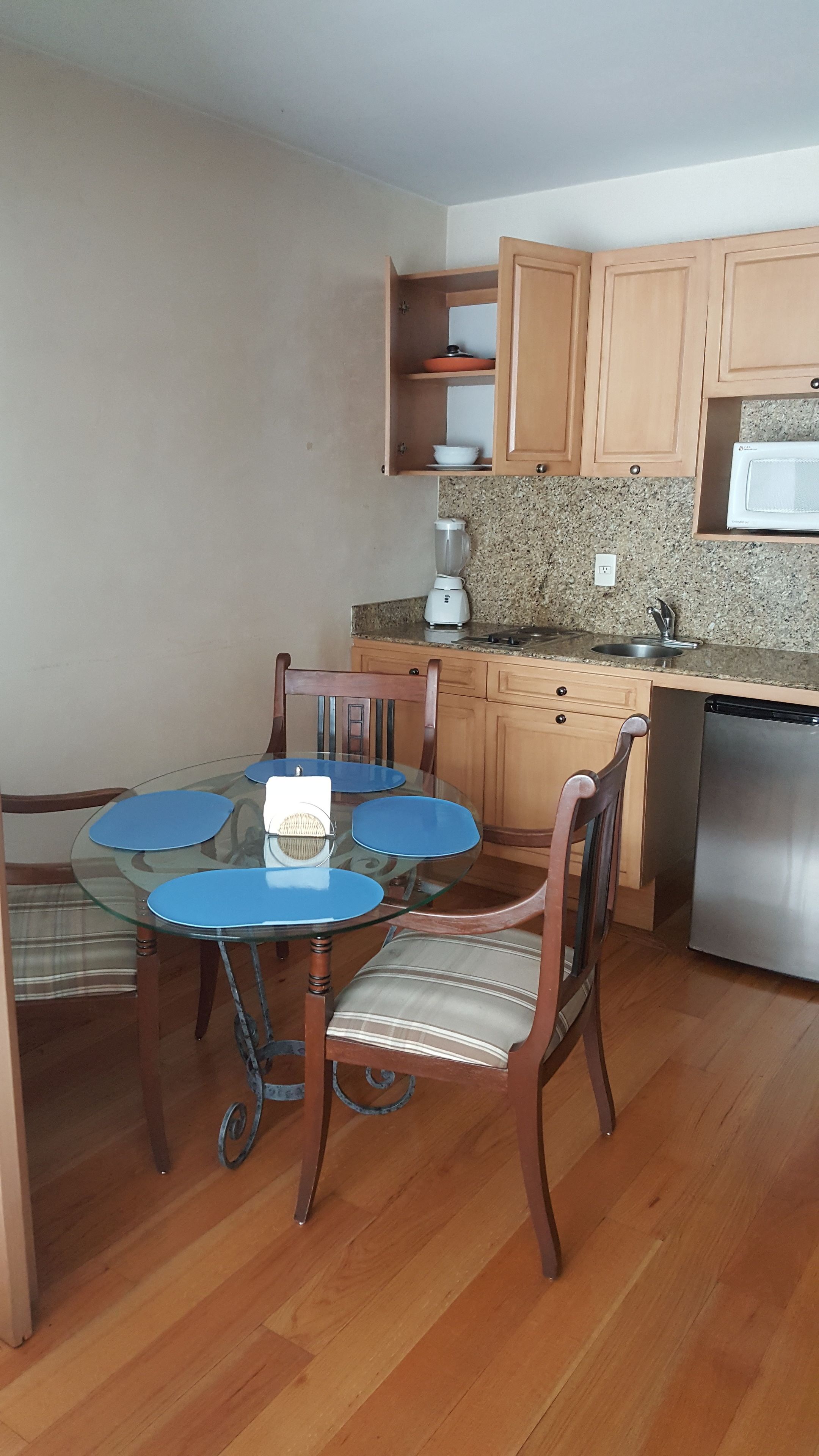 Una cocina compacta con gabinetes de madera, encimera de granito, microondas integrado y un comedor con mesa y sillas de vidrio.
