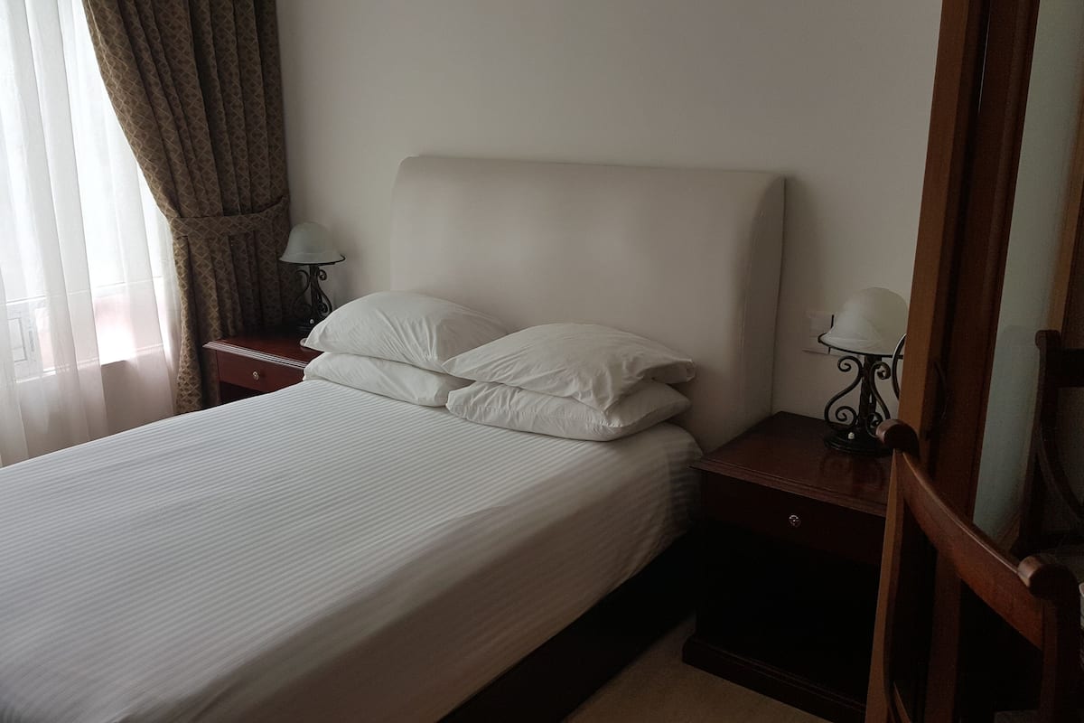 A neatly made bed with white linens, a wooden nightstand with a lamp, and a wooden chair with a table beside the bed.