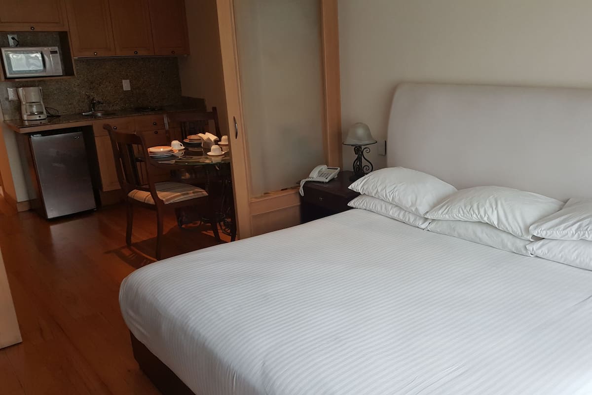 A hotel room with a large bed, a small kitchenette, a dining table with chairs, and a desk with a chair.