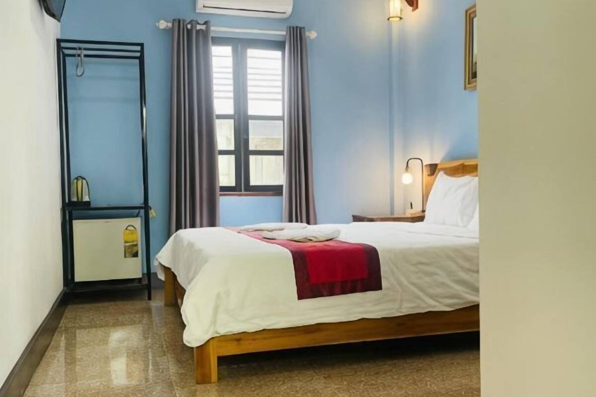 Quarto com cama, mesa de cabeceira, luminária, janela com cortinas e ar-condicionado fixado na parede.