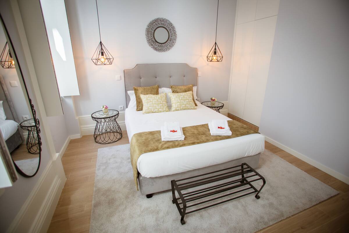 Une chambre bien rangée, avec un grand lit, des tables de chevet, un luminaire suspendu et un miroir décoratif accroché au mur.