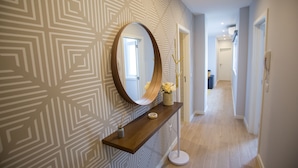 Un couloir avec un papier peint à motifs, un miroir rond, une console en bois sur laquelle sont posés un vase et un chandelier, et un téléphone fixé au mur.