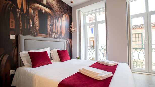 Une chambre d’hôtel avec un grand lit, une literie rouge et blanche, une fresque représentant des tonneaux de vin, et un balcon avec vue.