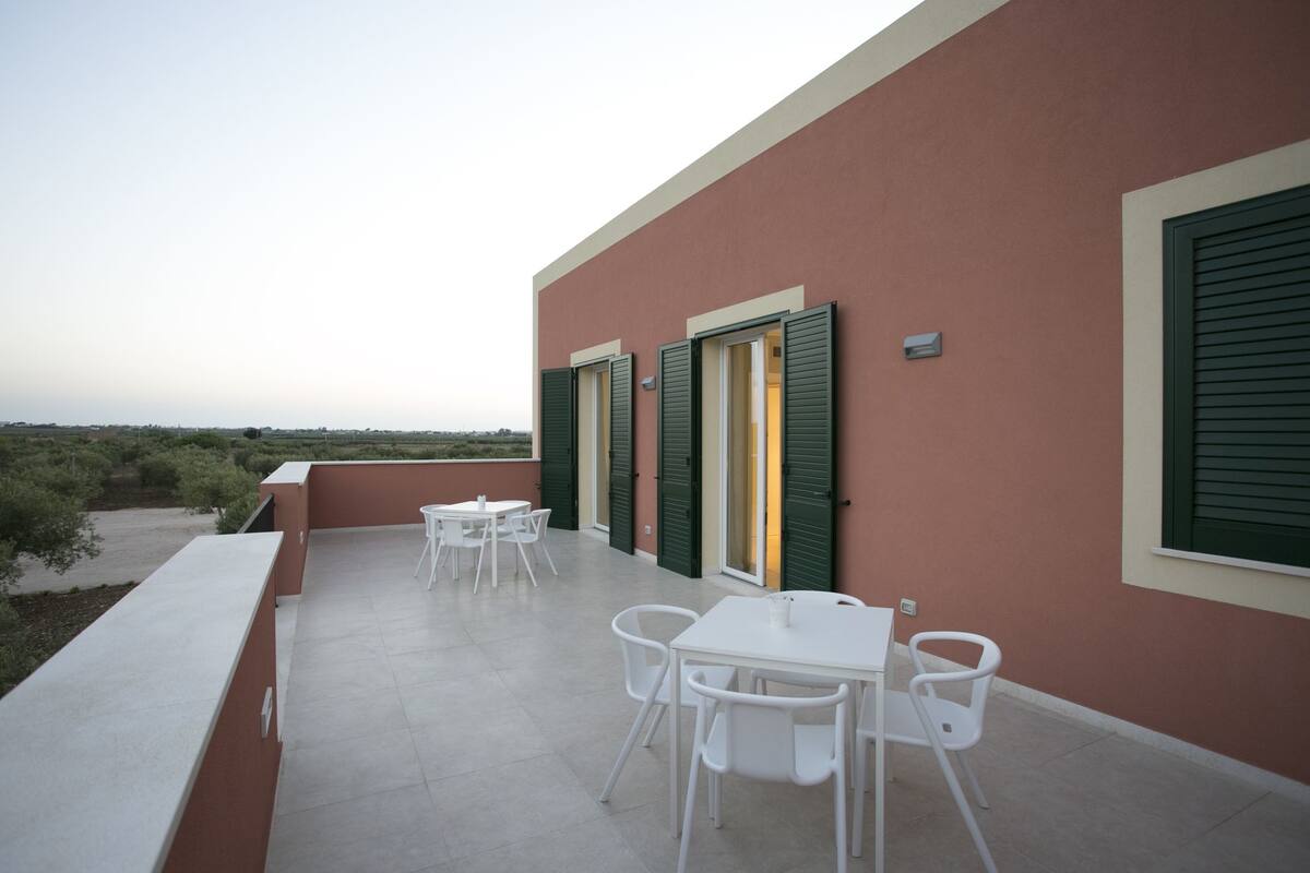 A terrace with white tables and chairs, green shutters, and a view of the landscape.