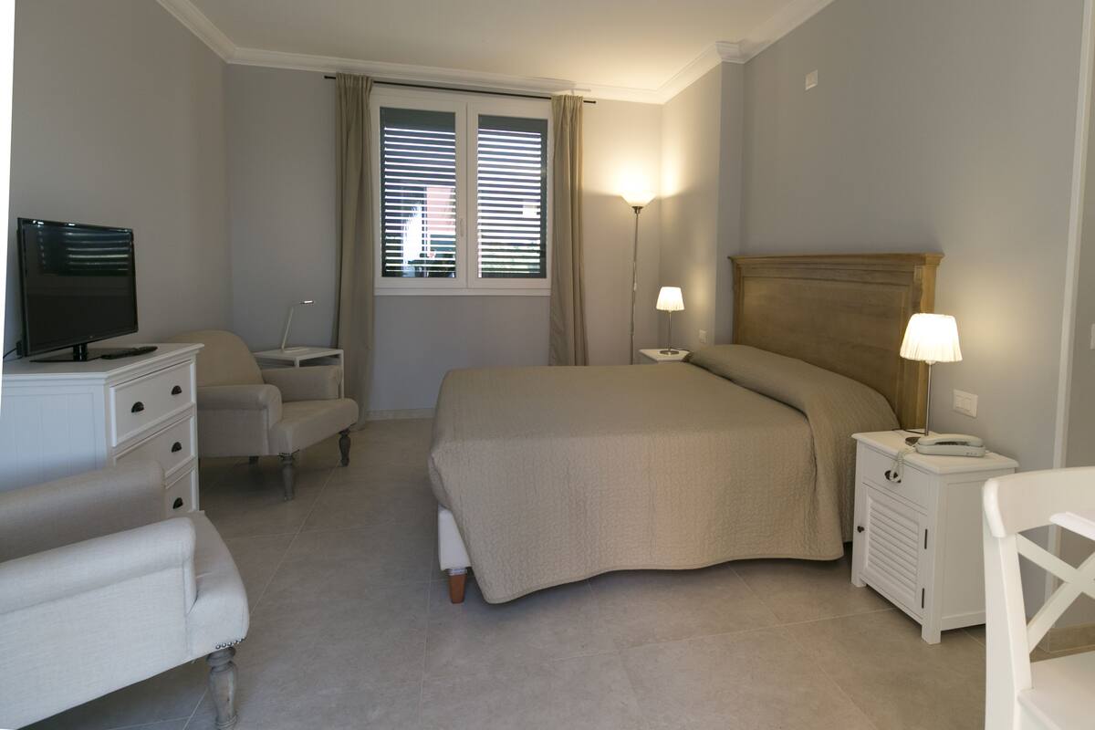 A bedroom with a bed, bedside tables, a desk, a chair, a television, and a window with blinds.