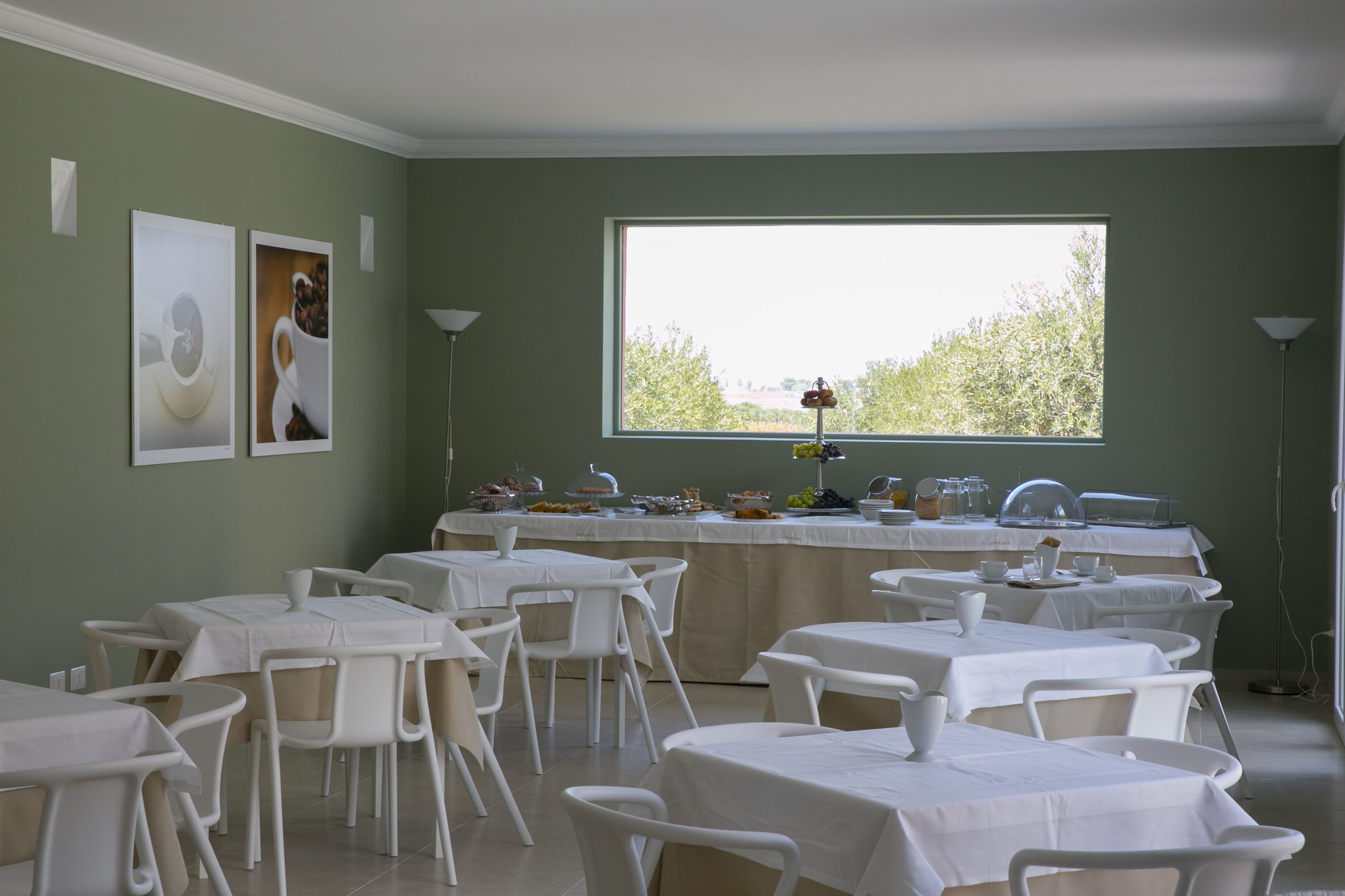 Un comedor con mesas y sillas, un bufé con comida y bebidas, y un amplio ventanal con vistas a árboles.