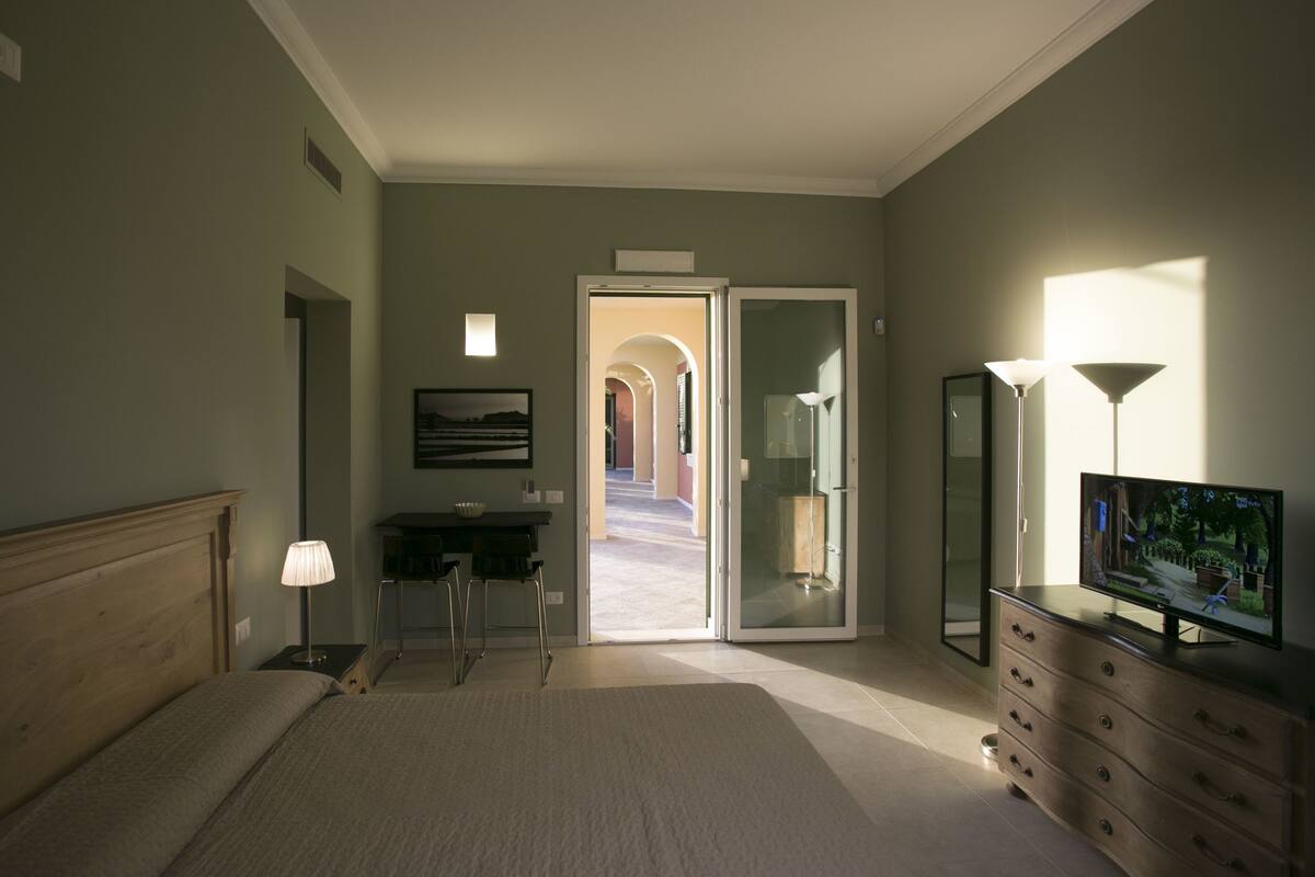 A bedroom with a bed, a desk, a dresser, a TV, and a door leading to a hallway.