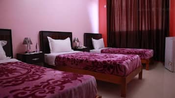 Habitación de hotel con tres camas, cada una con colcha estampada en rosa y morado. Hay dos mesitas de noche con lámparas y un refrigerador.