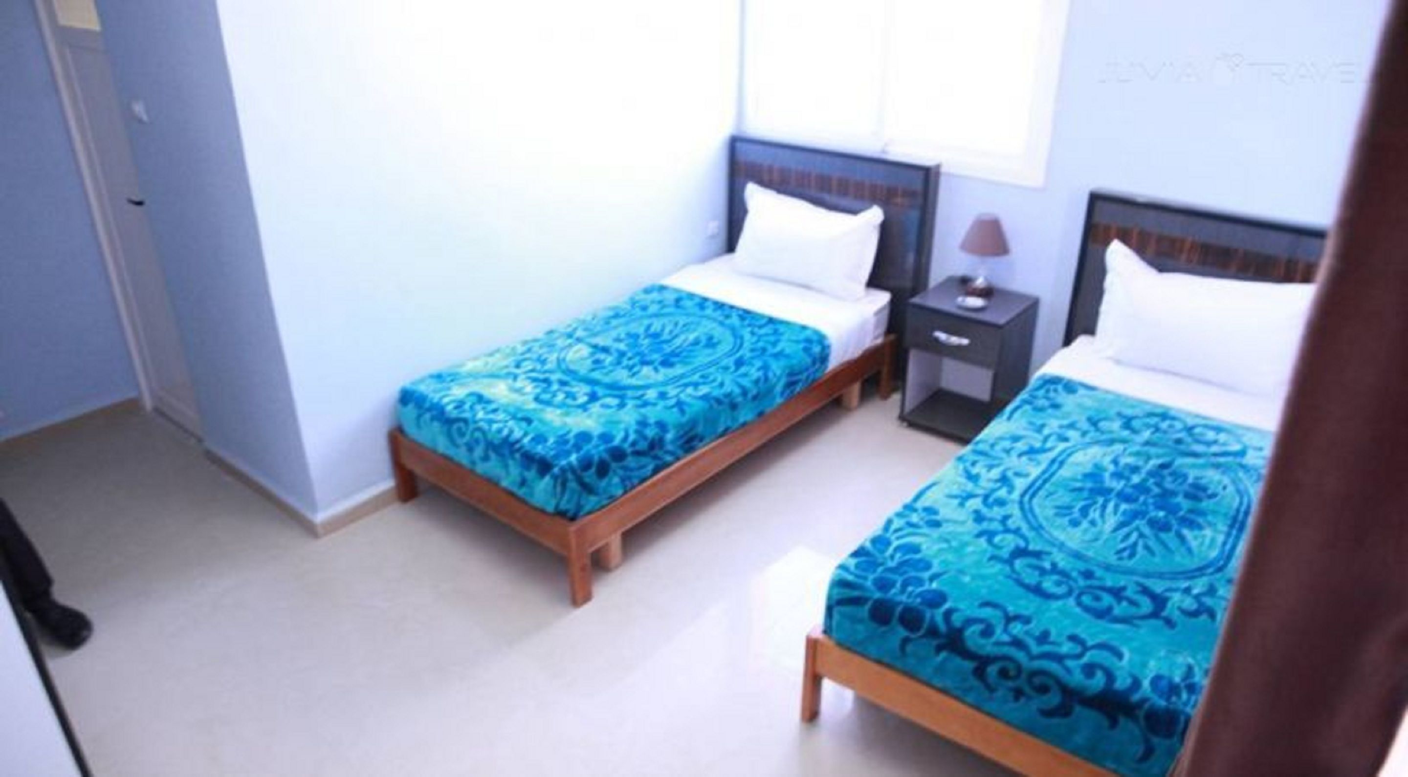 A bedroom with two single beds, each with a blue patterned bedspread and white pillows. There is a small bedside table with a lamp on one side and a drawer on the other. The room has light-colored walls and a door on the left side.