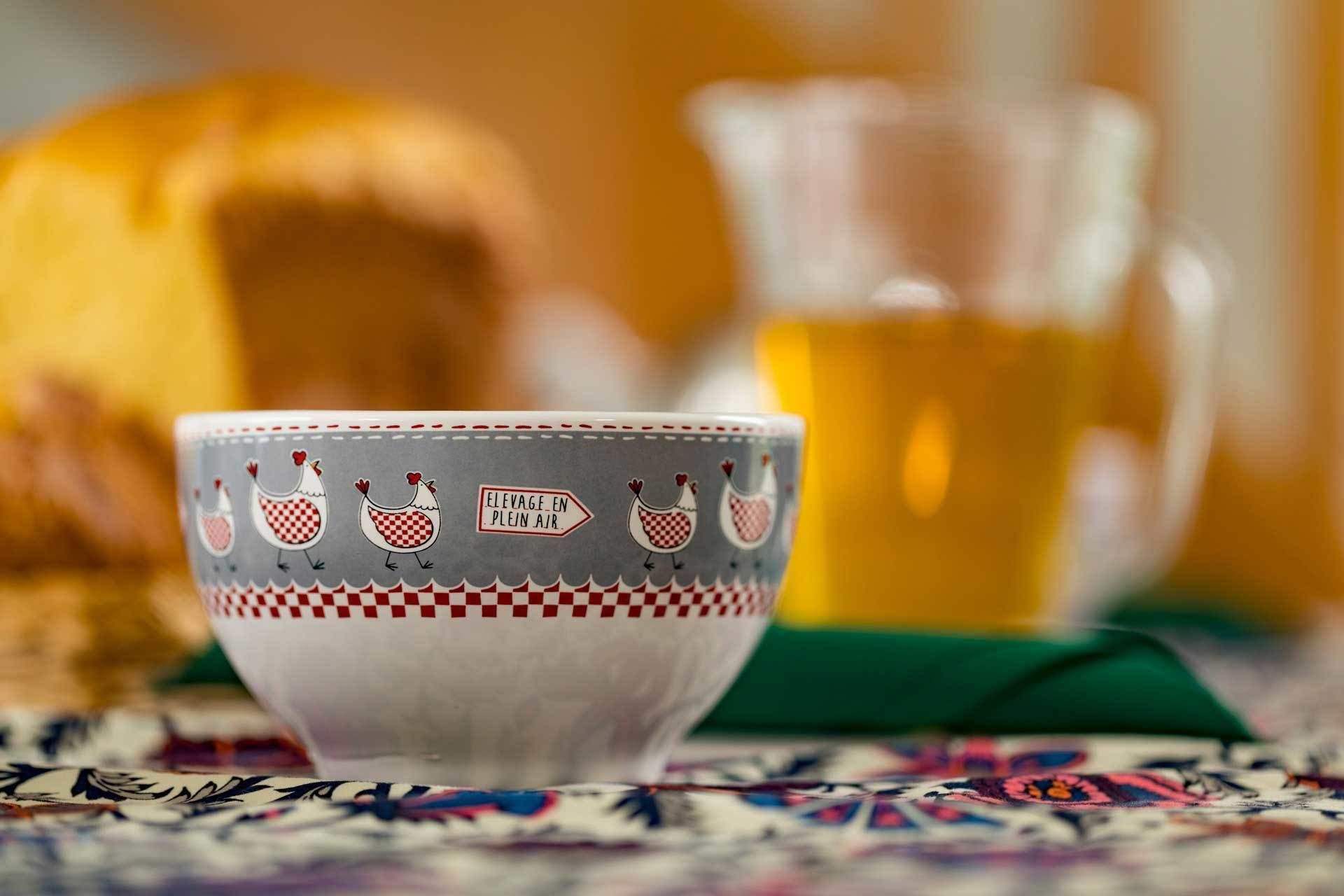 Un bol en céramique orné d’un motif à carreaux et de dessins de poulets, un pichet en verre et une miche de pain posés sur une nappe à motifs.