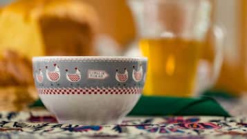 A ceramic bowl with a checkered pattern and chicken illustrations, a glass pitcher, and a loaf of bread on a patterned tablecloth.