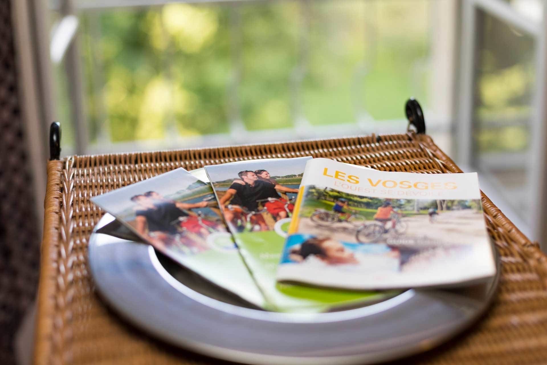 A wicker tray with a magazine titled "LES VOSGES" and a plate on a table near a window.