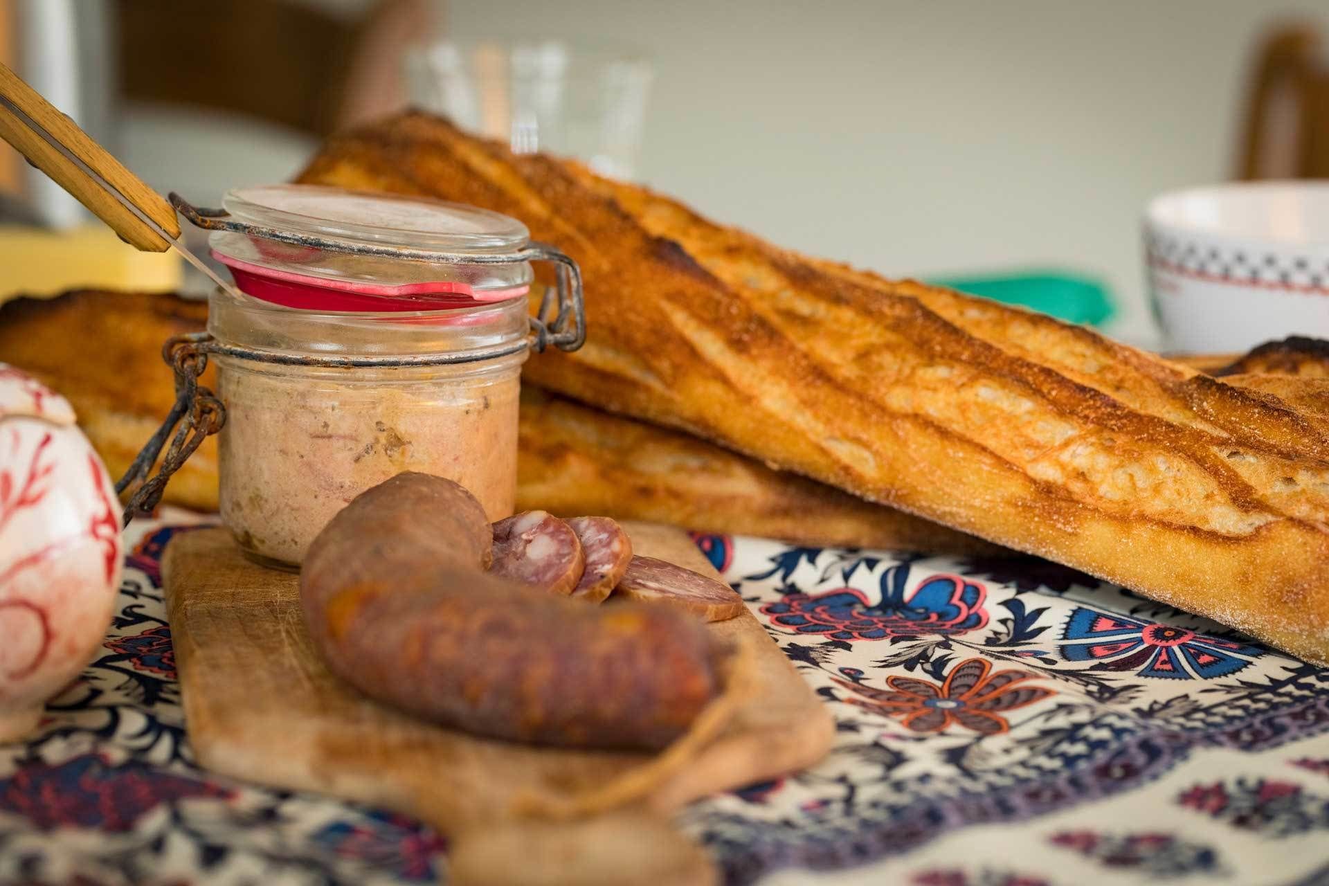 Een landelijke setting met een potje jam, een brood en een plak worst op een tafelkleed met dessin.