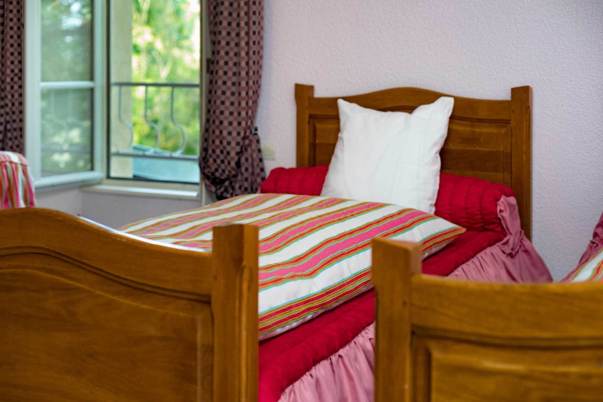 A wooden bed with a striped bedspread and red pillows in a room with a window and curtains.