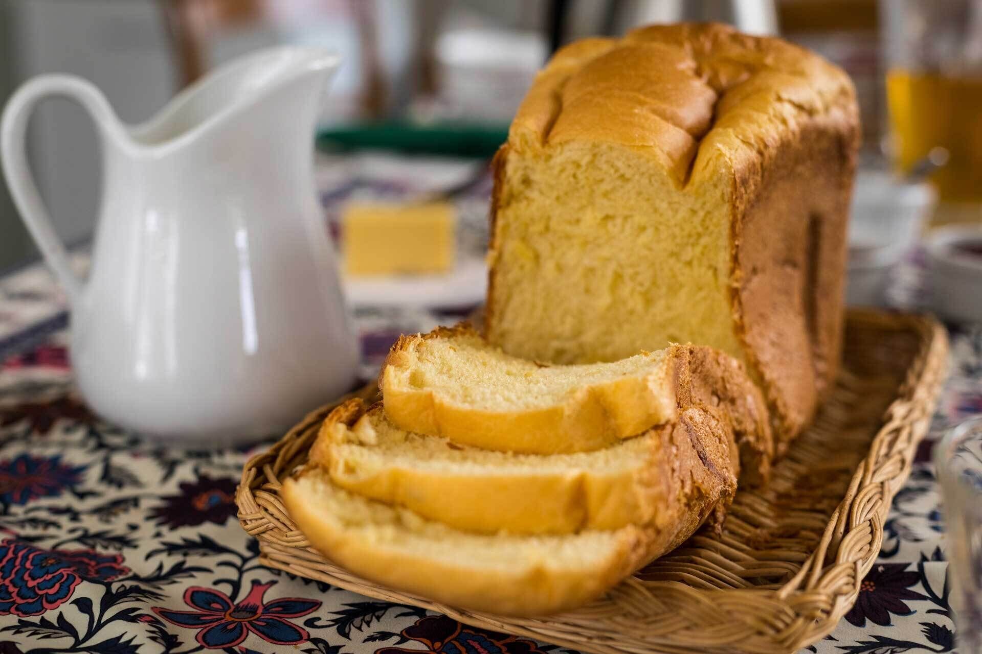 A loaf of bread on a woven basket, a white pitcher, and a patterned tablecloth.
