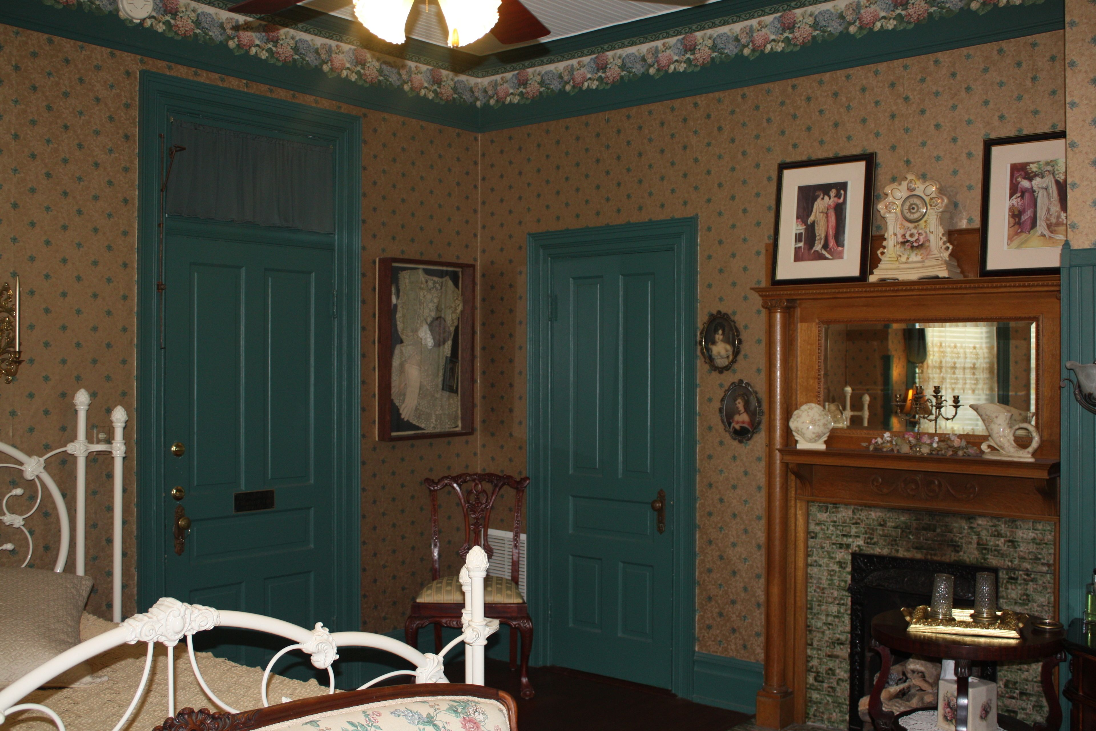 A room with two teal doors, a white metal bed frame, a wooden chair, a fireplace with a mirror above it, and floral wallpaper.