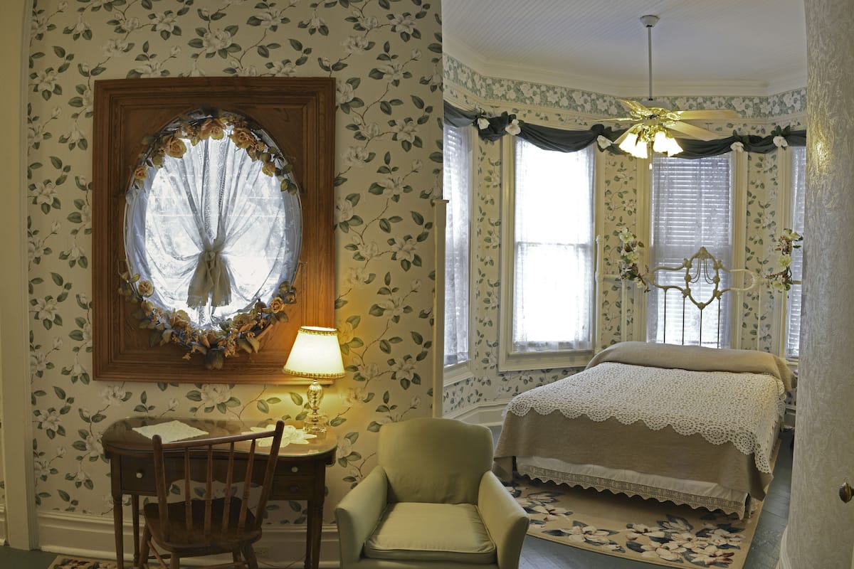 Une chambre avec un papier peint à motifs floraux, un lit recouvert d’une couvre-lit en dentelle, une chaise, un bureau avec une lampe et une fenêtre avec un rideau.
