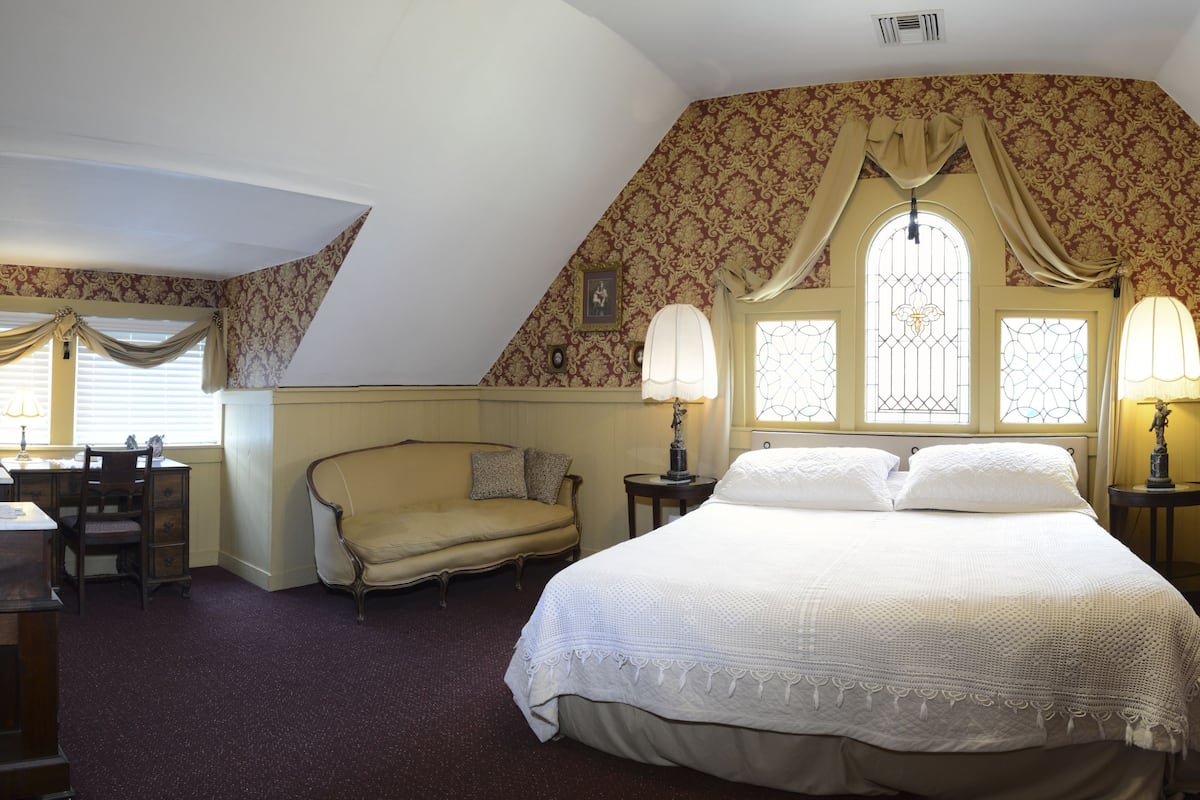 Une chambre avec un grand lit, deux tables de chevet avec des lampes, une chaise, un bureau et une fenêtre avec des rideaux.