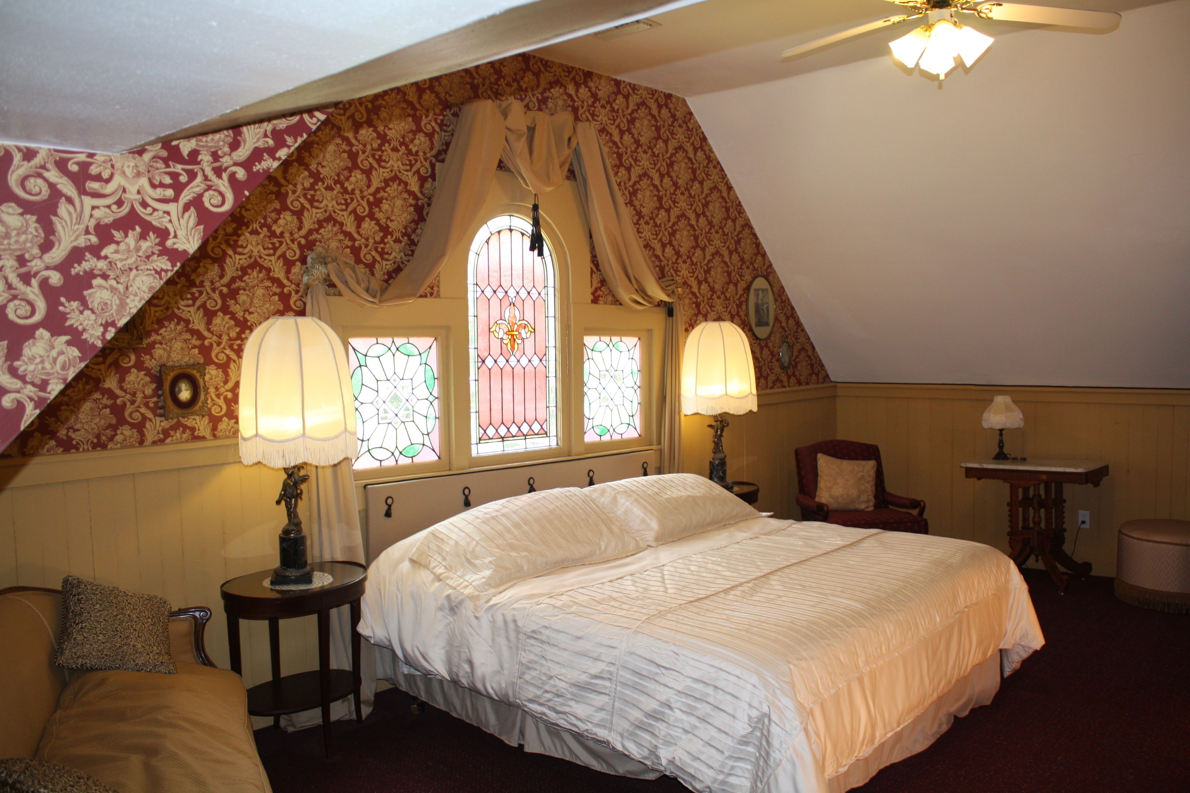 A bedroom with a large bed, two bedside lamps, a chair, a small table, and a window with stained glass.