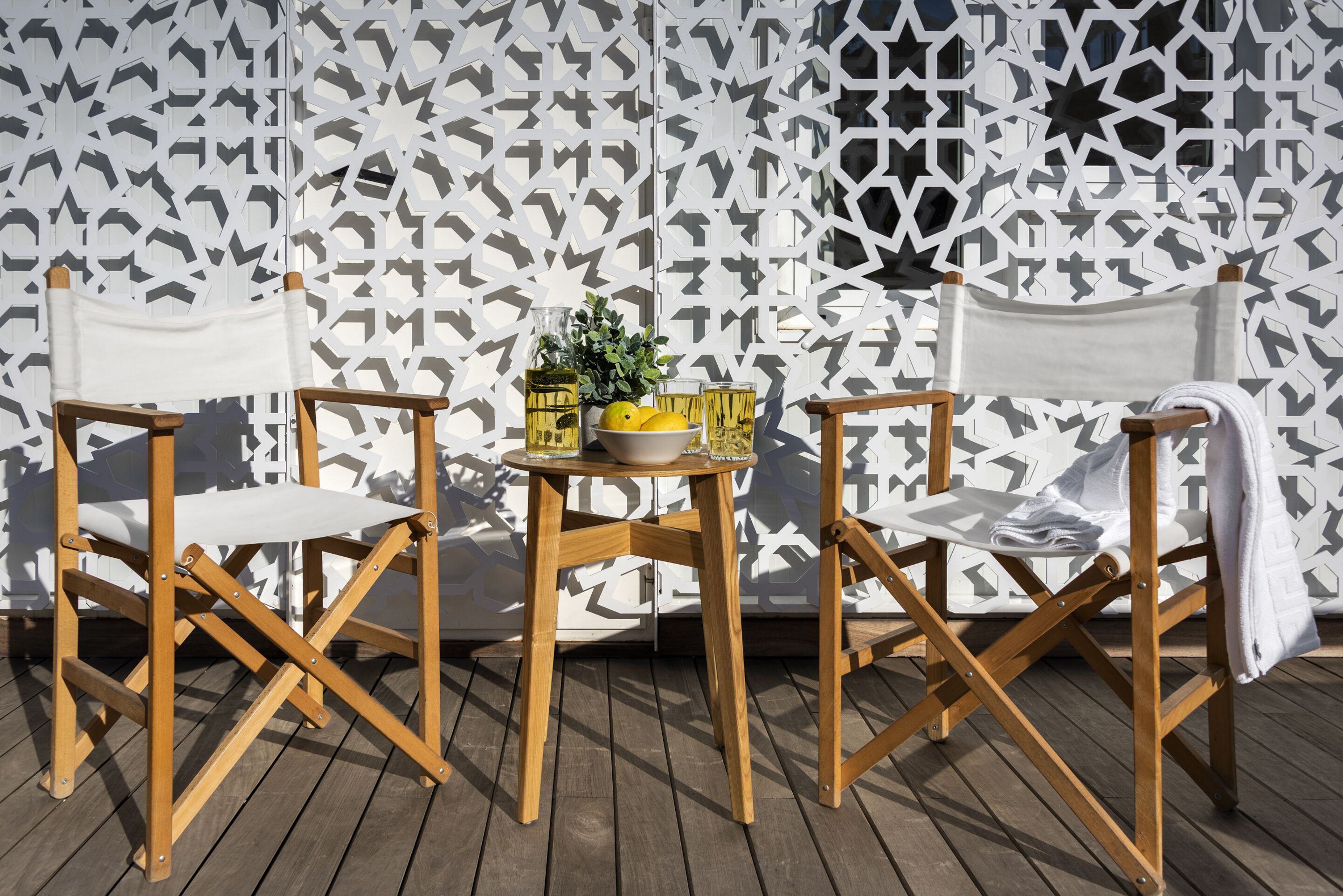 Una terraza de madera con dos sillas plegables y una pequeña mesa, frente a un muro con diseño.