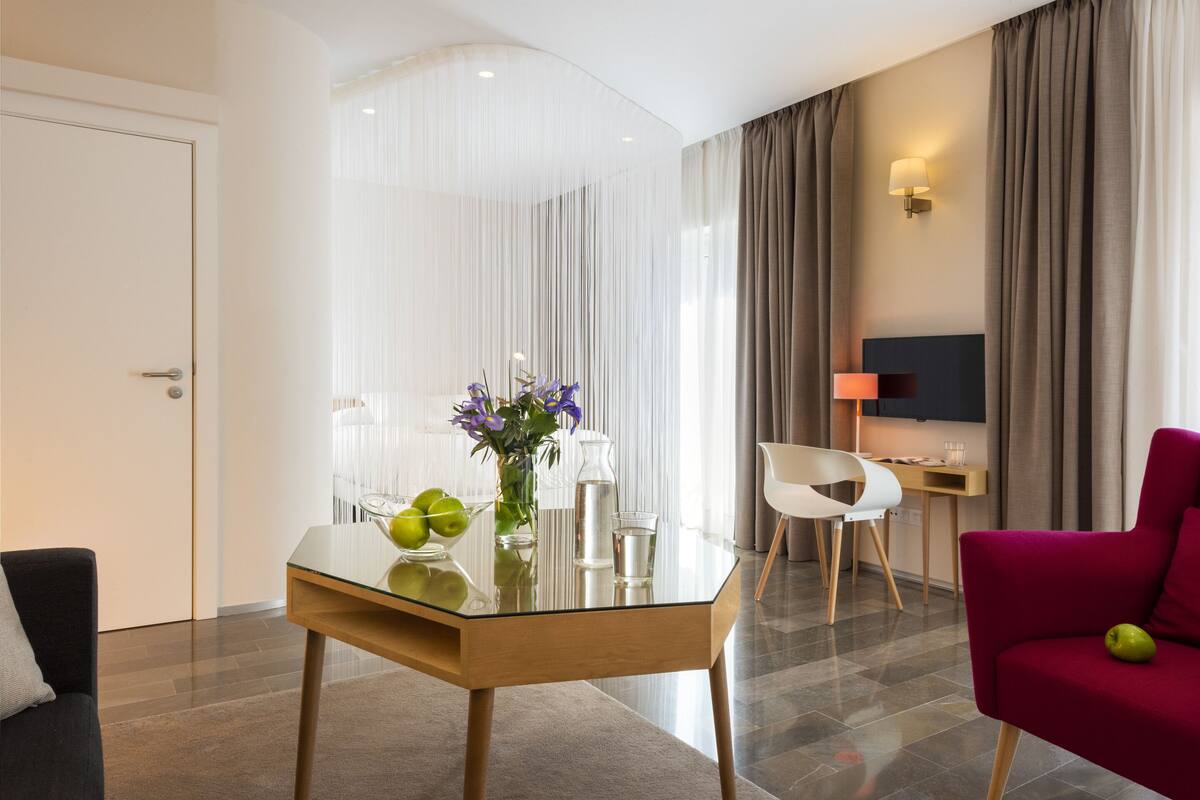 A modern hotel room with a glass dining table, a red sofa, a TV, and a view of the city.