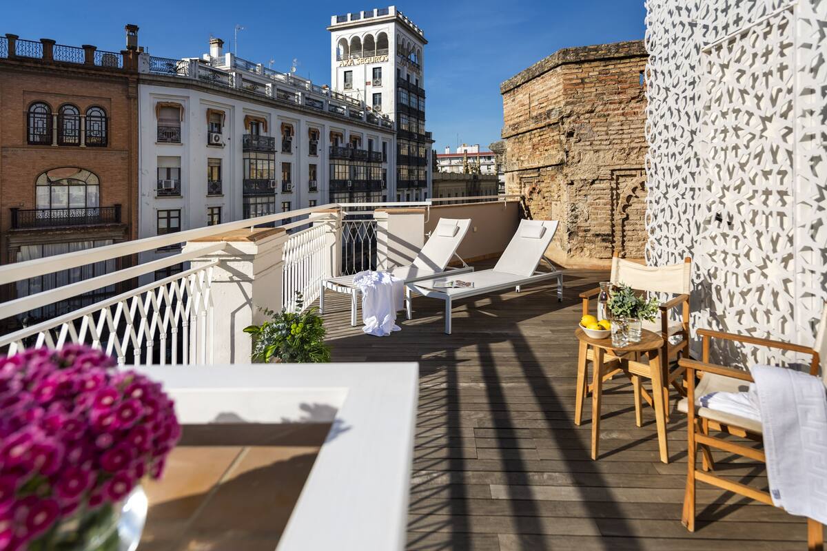 A rooftop terrace with lounge chairs, a table with a vase of flowers, and a view of historic buildings.