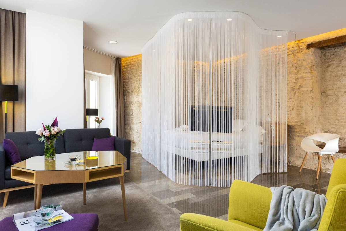 A modern hotel room with a glass partition, a sofa, a dining table, and a yellow armchair.