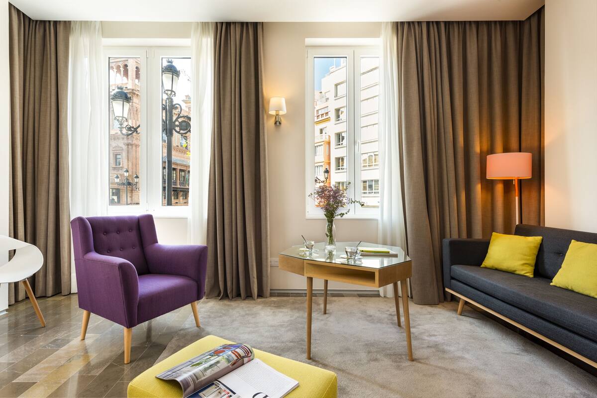 A modern living room with a purple armchair, a sofa with yellow cushions, a glass coffee table, and a view of a cityscape through large windows.
