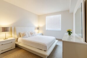 A bedroom with a bed, bedside tables, a nightstand, a window with blinds, and a mirror.