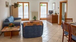 Ein Wohnzimmer mit einer Couch, einem Couchtisch und einem Essbereich mit Stühlen und einem Tisch. Es gibt zwei Fenster mit Vorhängen, eine Heizung und ein Fernseher auf einem Holzschrank.