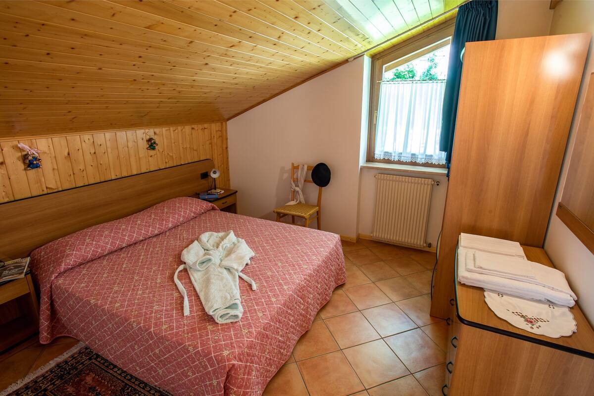 Ein Schlafzimmer mit Holzboden, einem Bett mit roter Bettdecke, einem Nachttisch aus Holz, einem Stuhl, einem Spiegel und einem Fenster mit Vorhängen.