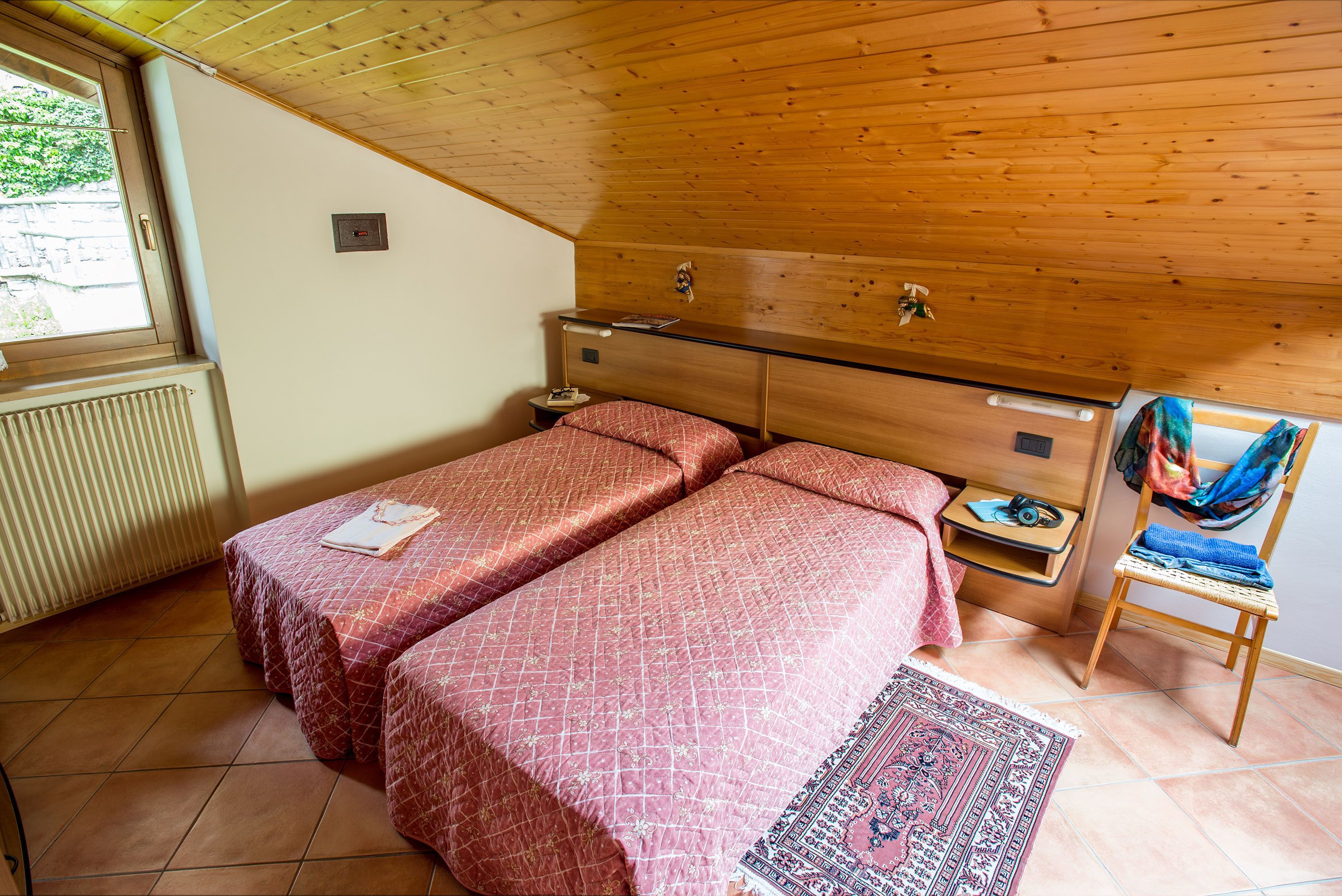 A bedroom with two beds, a wooden headboard, a small bedside table, a chair, and a window.