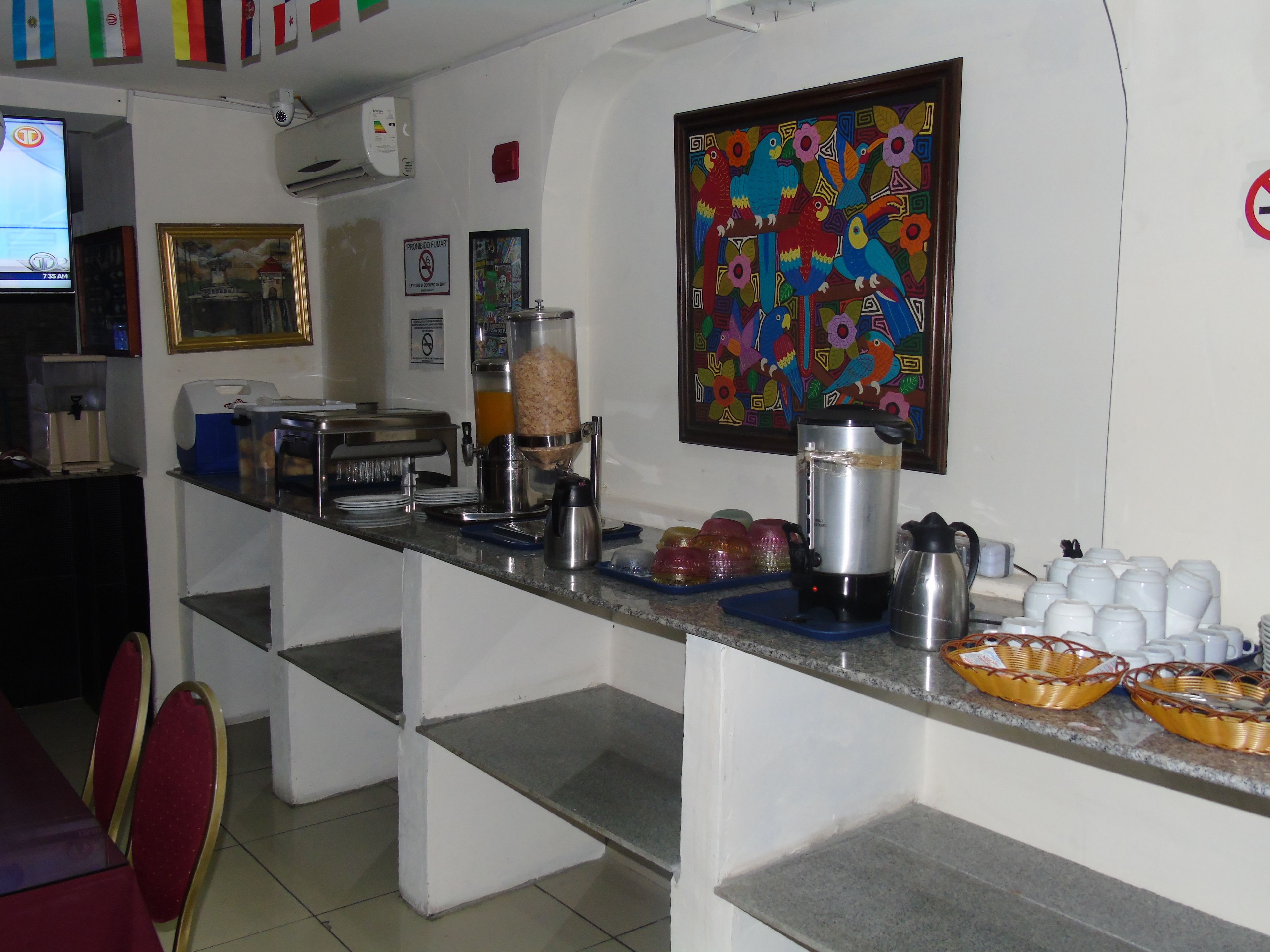 A breakfast bar with a coffee machine, juice dispenser, and various food items. There are chairs and a television in the background.