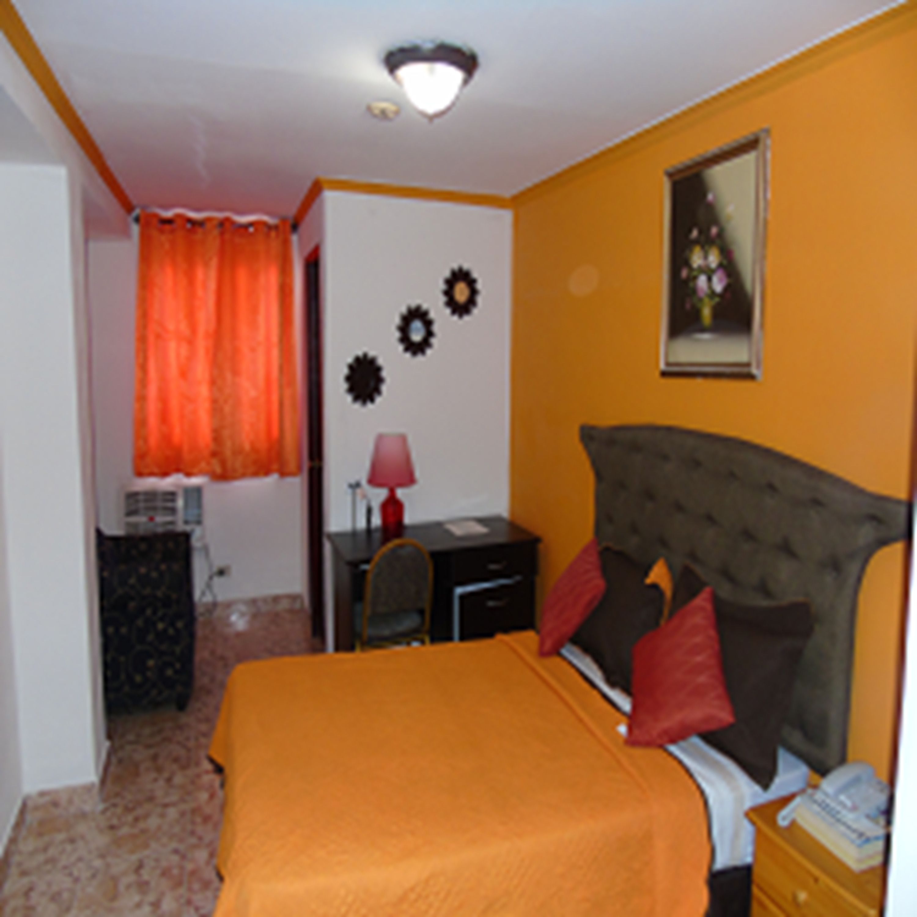 Un dormitorio con colcha naranja, un escritorio con lámpara, una silla y un aparador.