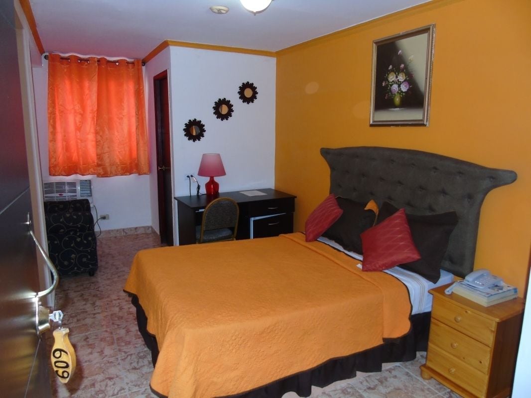 A bedroom with a bed, a nightstand, a desk, a chair, a lamp, and a door.