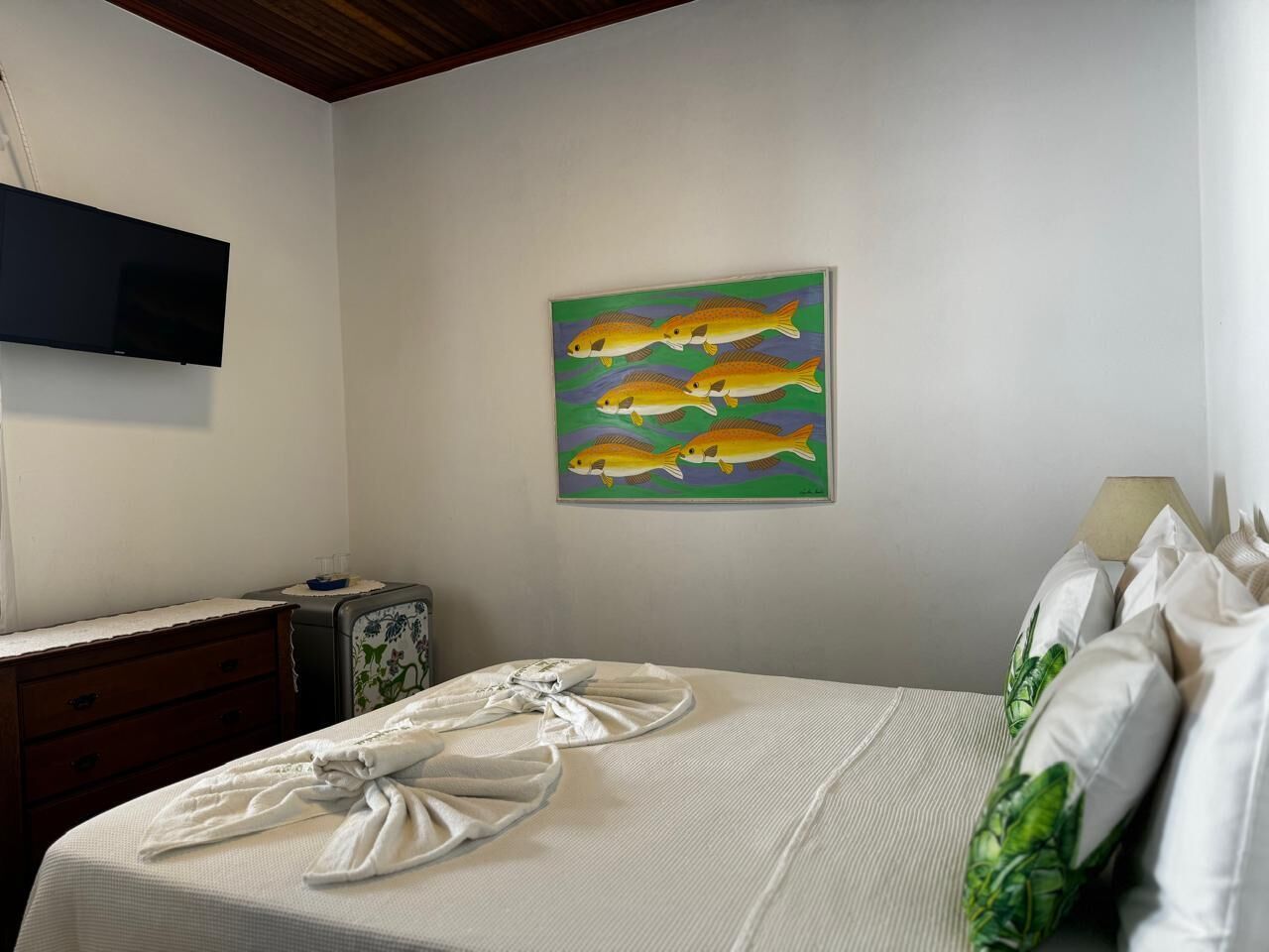 Una cama bien hecha con toallas, un cuadro de peces en la pared, un televisor y una lámpara.