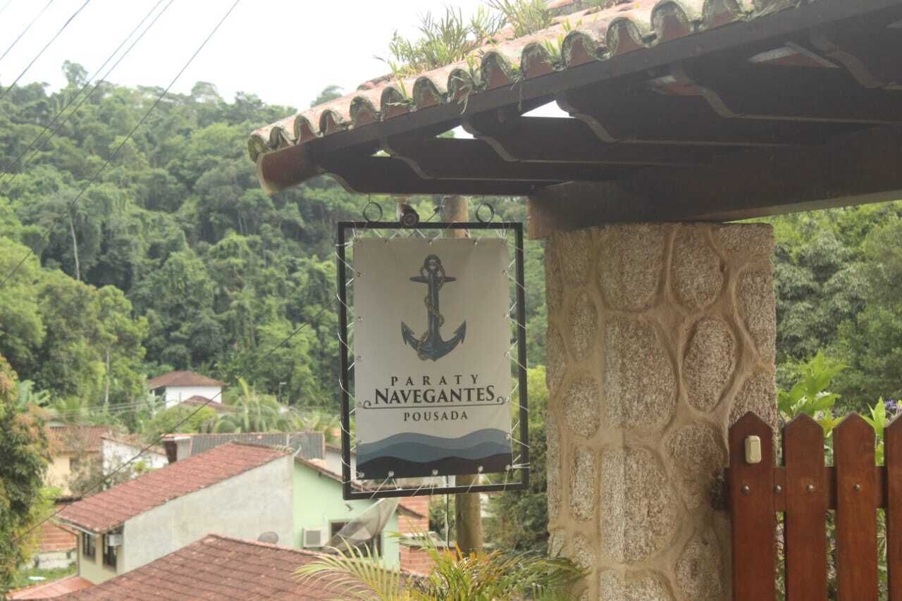 A sign for 'Paraty Navegantes, Pousada' with an anchor symbol, set against a backdrop of lush greenery and residential buildings.