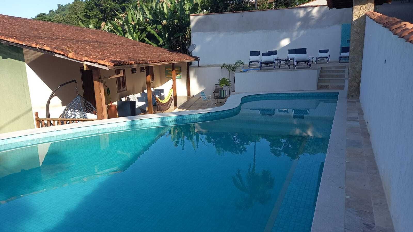 A swimming pool with a tiled deck, a covered seating area with chairs, and a white wall with a window.