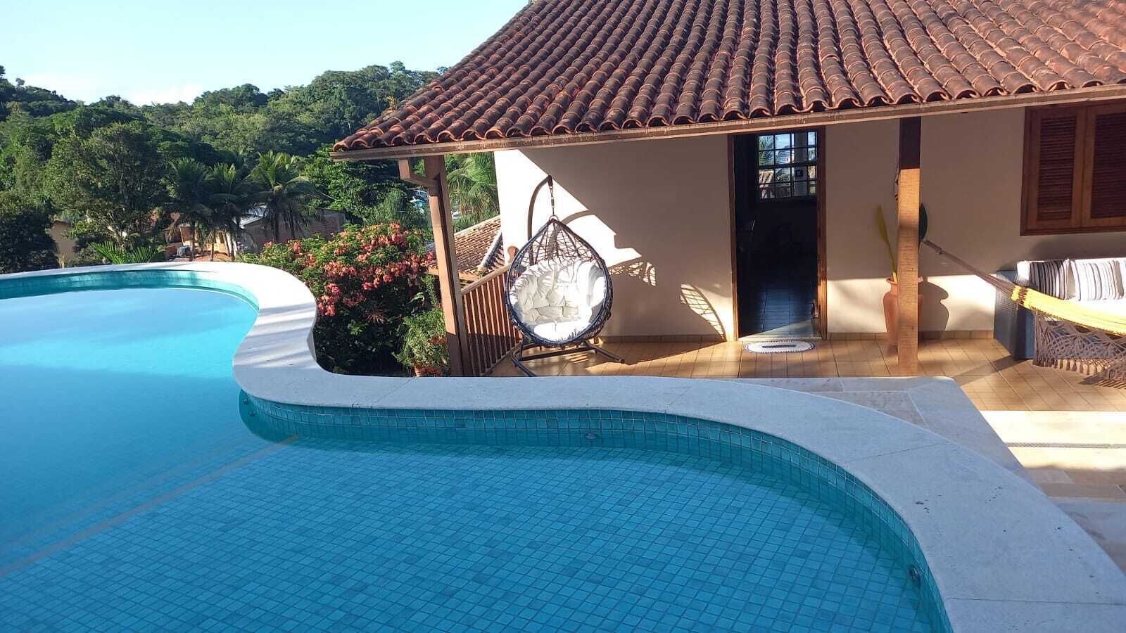 Ein Pool mit einer Fliesenbordsteinkante, ein überdachter Sitzbereich mit einem Stuhl und ein Gebäude mit einem Flachdach.