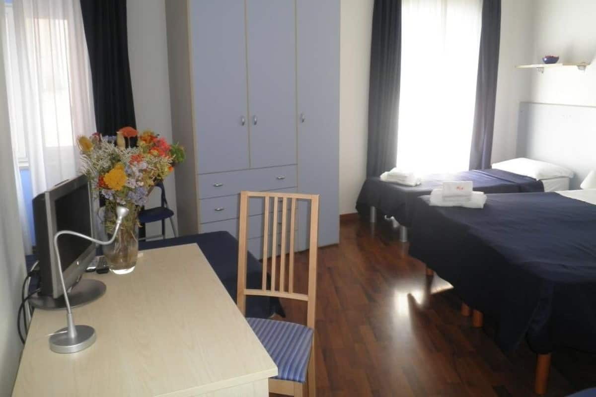 A hotel room with a desk, chair, wardrobe, two beds, and a bedside table.