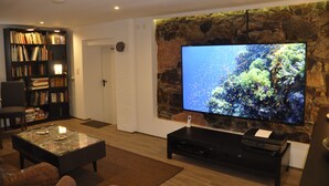A living room with a stone wall, a large flat-screen TV, a bookshelf, and a coffee table. - Pension Schwedenkreuz (Reimerath)