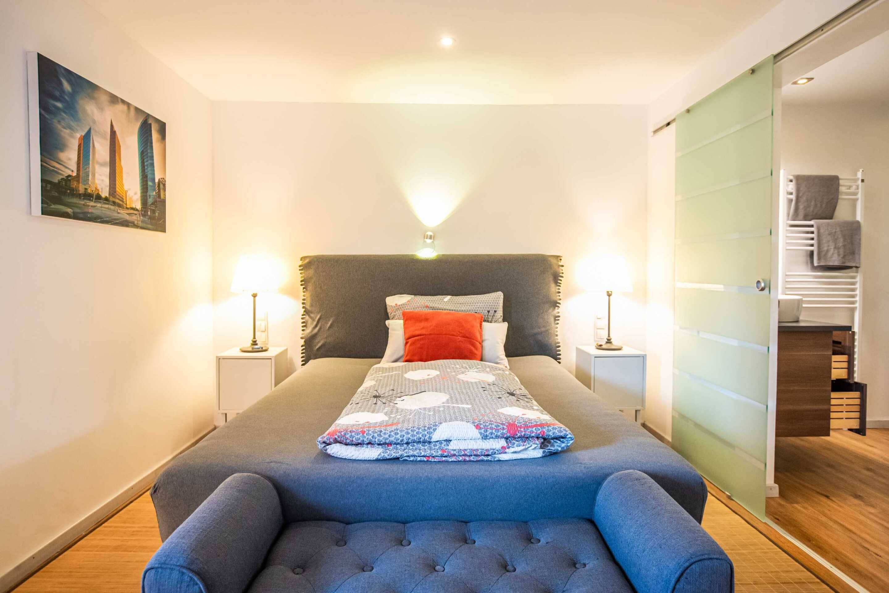 Un dormitorio con cama, lámparas de noche, una mesita de noche, una silla, un cuadro en la pared y una puerta corredera que da a un baño.