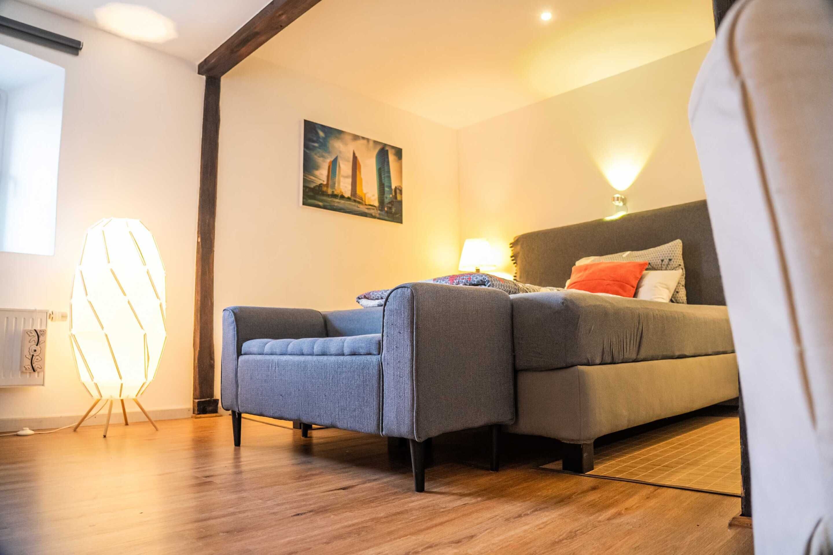 Un dormitorio moderno con un sofá gris, una mesita de noche con una lámpara y un cuadro en la pared.