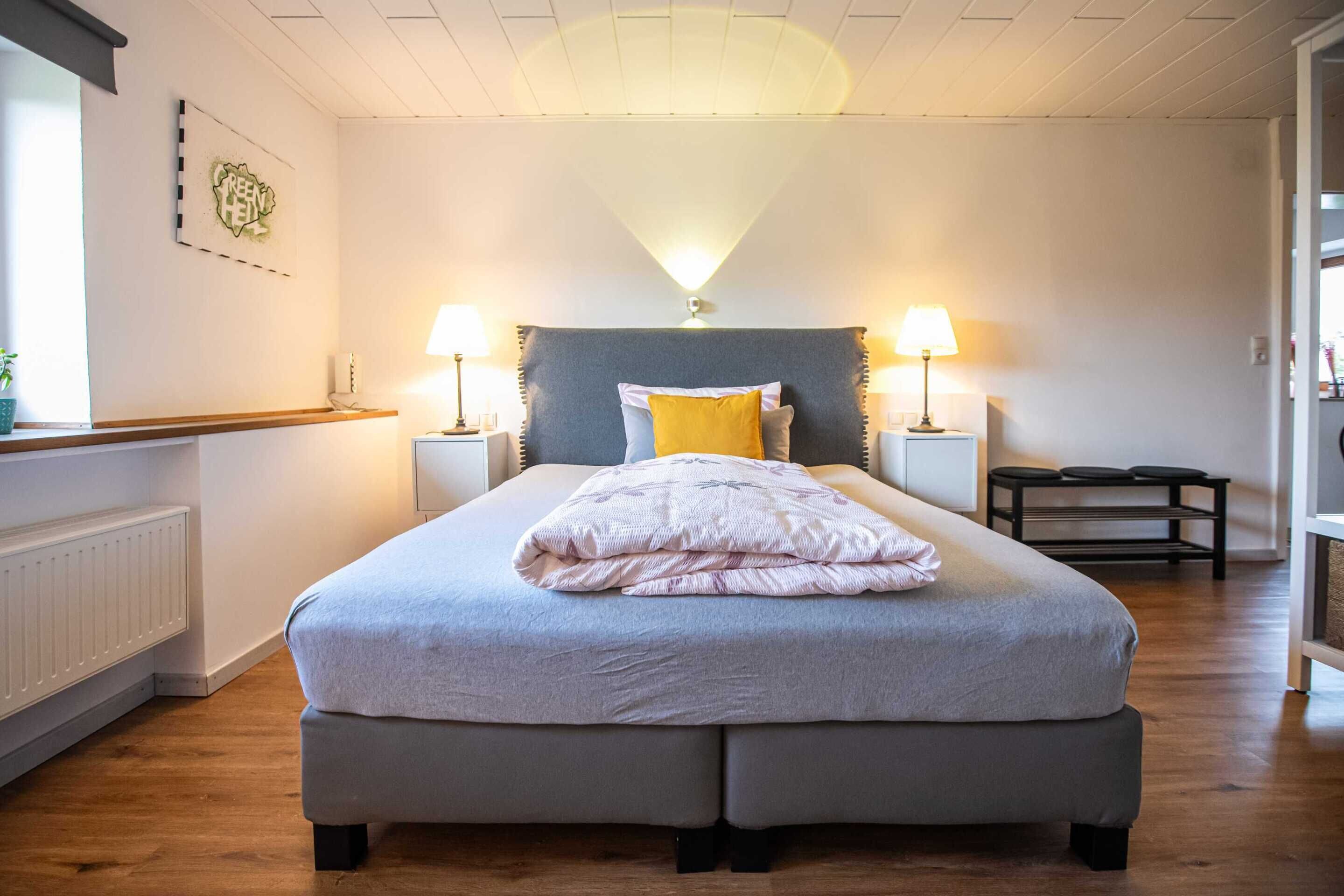 A bedroom with a bed, two bedside lamps, a nightstand, a window with a plant, and a radiator.