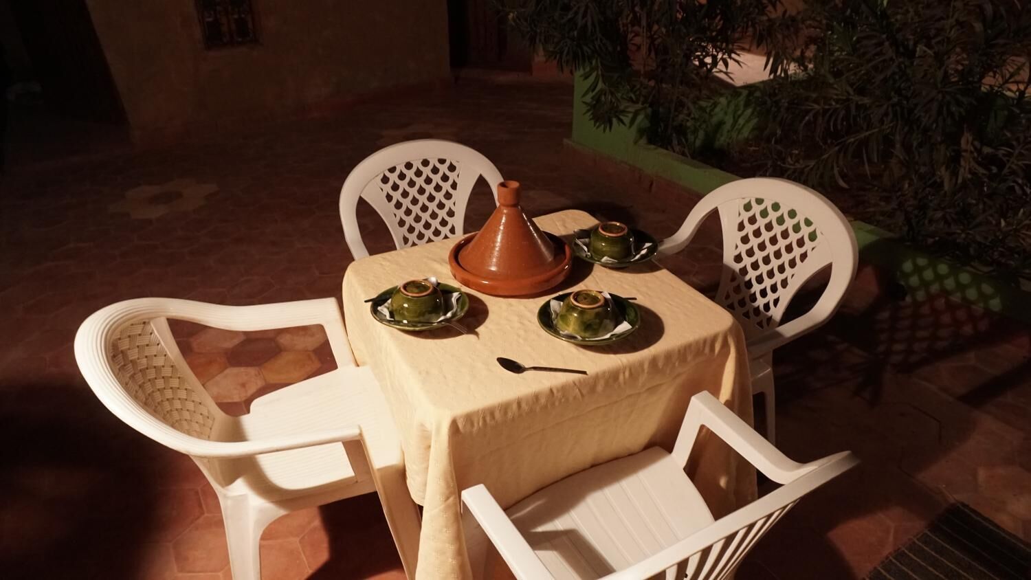 Una mesa dispuesta con un tajín marroquí, un hervidor de té y tazas, rodeada de sillas blancas en un entorno exterior.