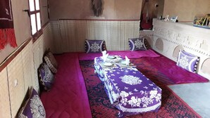 Een klassieke kamer met een bed, kussens, een tafel met tafelkleed, een gitaar en een aan de muur bevestigd kruis.