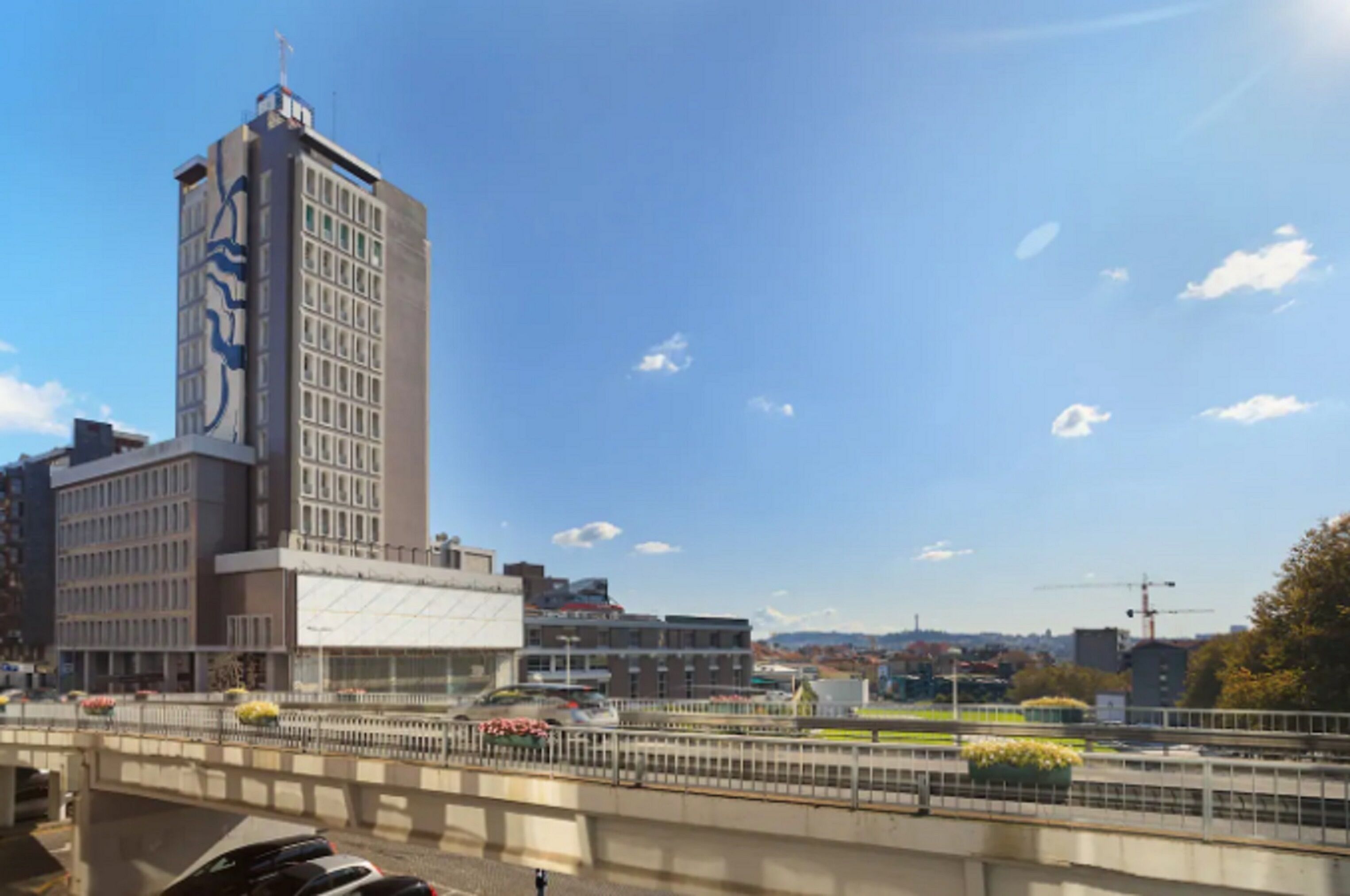 A cityscape with a prominent building featuring a large blue design on its facade, a bridge with a metal railing, and a clear blue sky with scattered clouds.