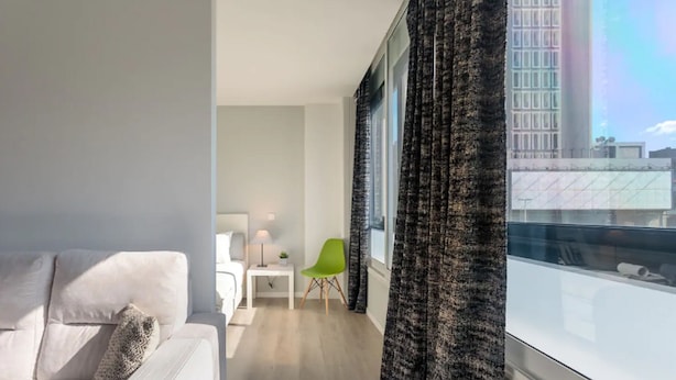 A modern living room with a white sofa, a green chair, a small table with a lamp, and a view of a cityscape through large windows with dark curtains. - Downtown Porto Spot - 2 (Porto)