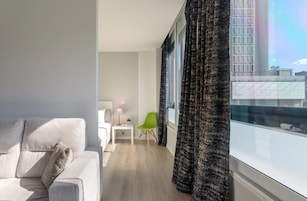 Uma sala de estar moderna com um sofá branco, uma poltrona verde, uma mesinha com uma luminária e vista para um horizonte urbano através de janelas amplas com cortinas escuras.